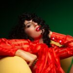 Low angle of confident sensual female with red lips touching hair sitting on chair and looking at camera