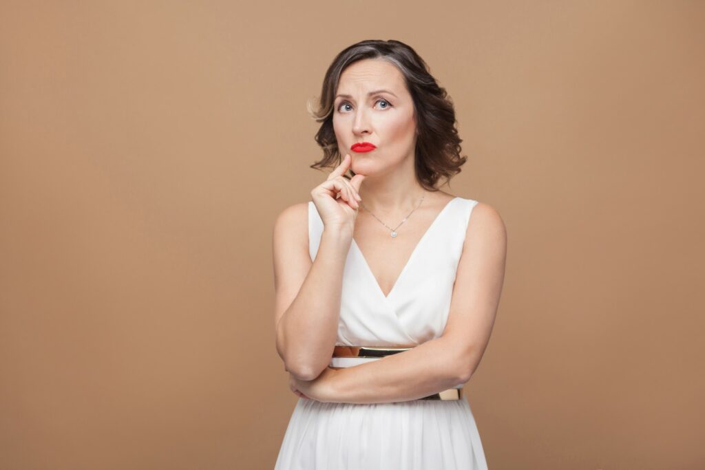 handsome middle aged woman thinking puzzled emotional expressing woman white dress red lips dark curly hairstyle studio shot indoor isolated beige light brown background