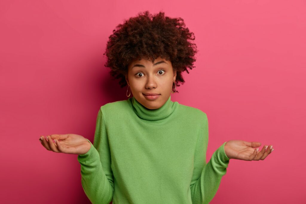 indifferent confused black woman spreads palms confusingly has doubts wears green turtleneck has hesitant face expression isolated pink space