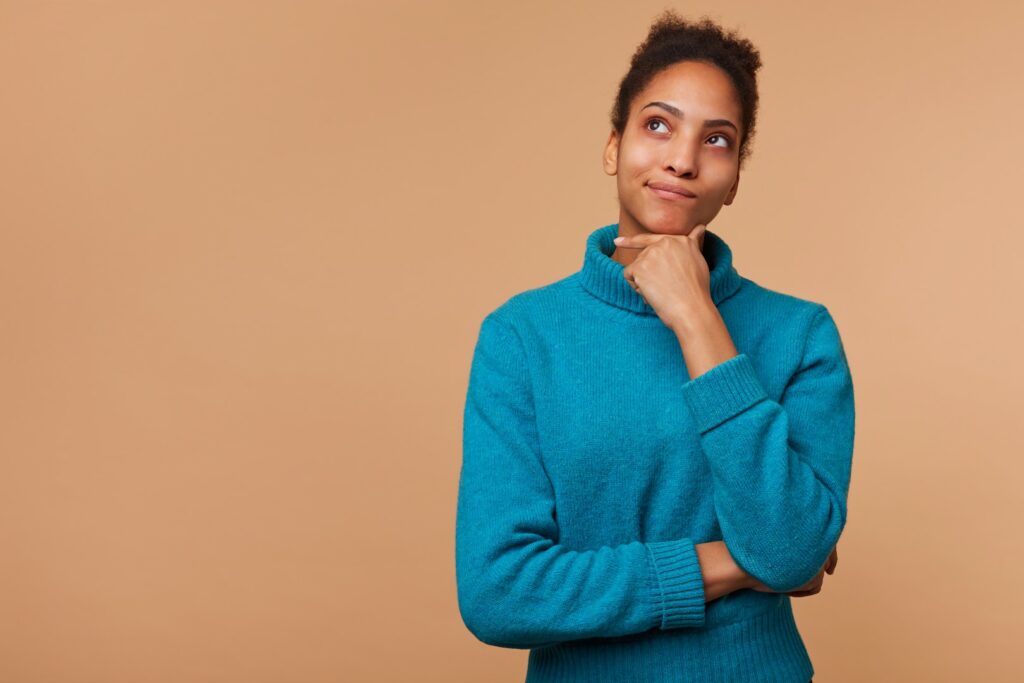 photo puzzled african american girl with curly dark hair wearing blue sweater touches chin can t decide doubts look up isolated beige background with copy space